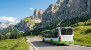 Autobus di linea su strada di montagna nelle Dolomiti, tra prati verdi e pareti rocciose sotto cielo sereno