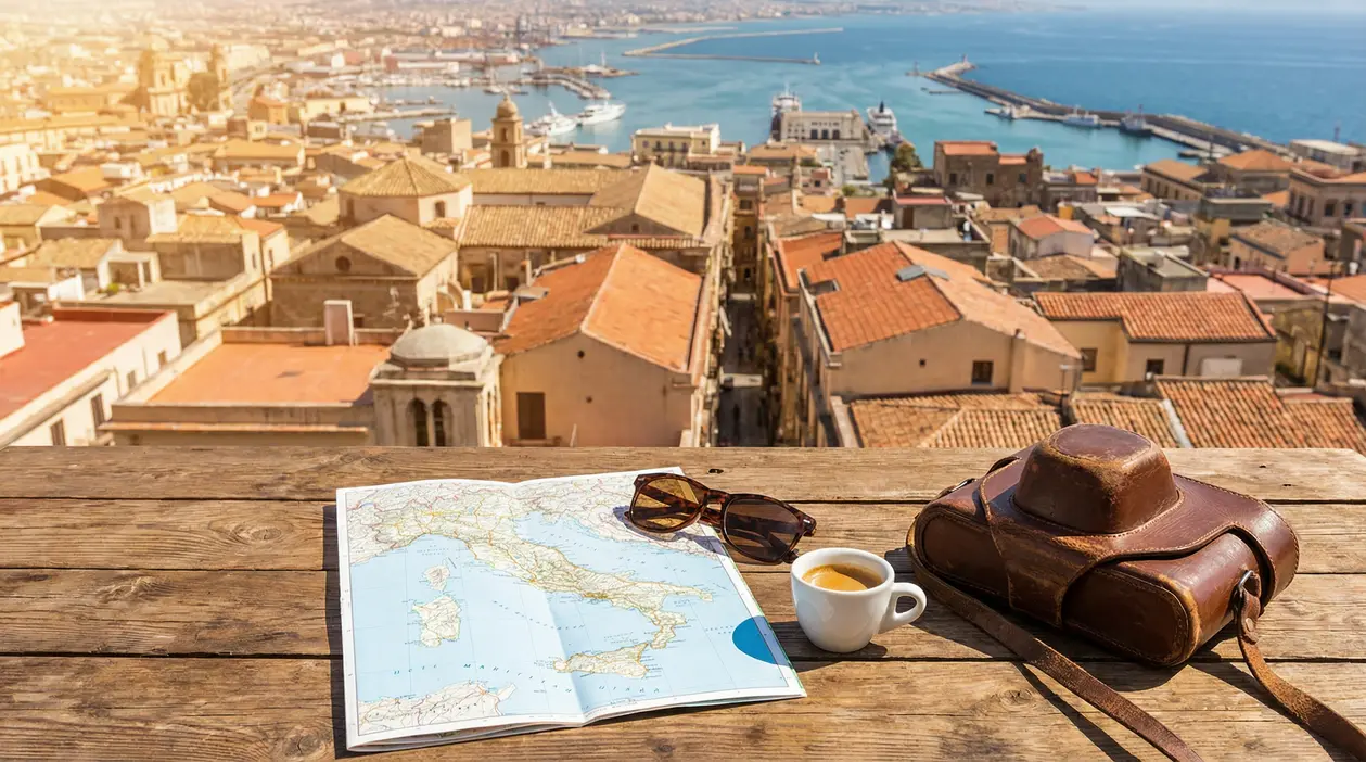 Mappa dell’Italia, occhiali da sole, caffè e borsa su un tavolo con vista su città costiera e porto