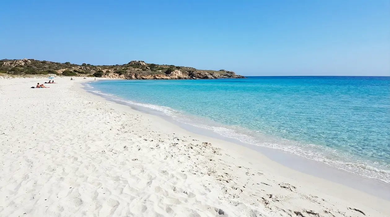 Spiaggia di sabbia bianca con mare turchese e costa rocciosa, con alcune persone in lontananza