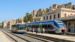 Treno regionale e autobus in stazione ferroviaria in Sicilia, con viaggiatori in banchina e borgo sullo sfondo